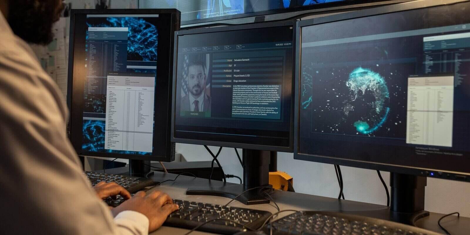 policeman entering personal data of suspect into computer database while sitting in front of monitors in office