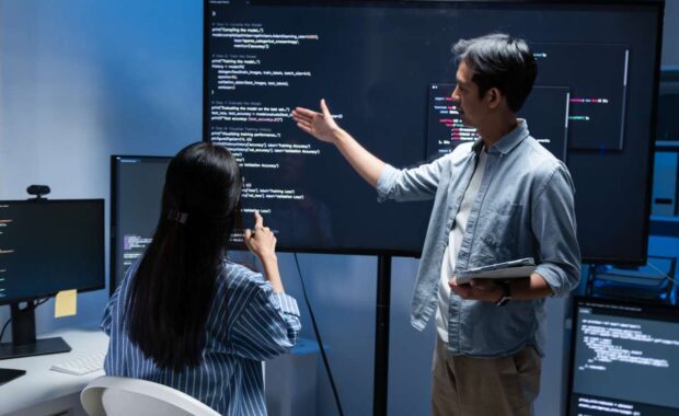 Software Developer Team working late in office at night. Programmer team collaboration, working on coding task, reviewing code while Male Programmer presenting code in the screen in tech office.
