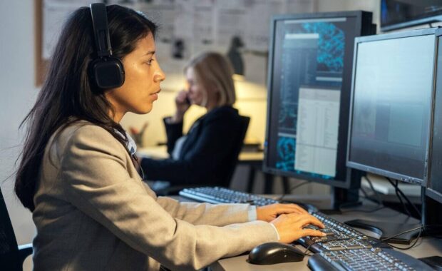 female operator of surveillance system sitting by desk in front of computer monitors in office