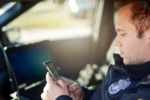 Police, officer and man texting with phone in patrol car for security contact, law enforcement update and mobile notification. Policeman typing message on cellphone for crime news, connection and app.