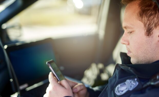 Police, officer and man texting with phone in patrol car for security contact, law enforcement update and mobile notification. Policeman typing message on cellphone for crime news, connection and app.